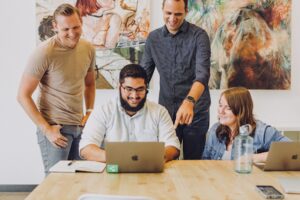Four people smiling, looking at a laptop on a wooden table, discussing enterprise search